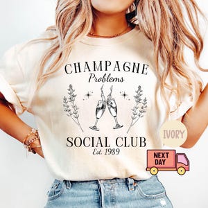 May include: A white t-shirt with the text "Champagne Problems Social Club Est. 1989" and a graphic of two champagne glasses clinking. The shirt is available in ivory.