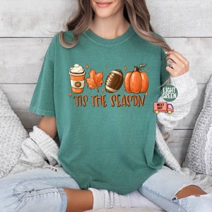 May include: A teal green t-shirt with a graphic of a coffee cup, a football, a pumpkin, and fall leaves. The text "'Tis the season" is printed on the shirt.