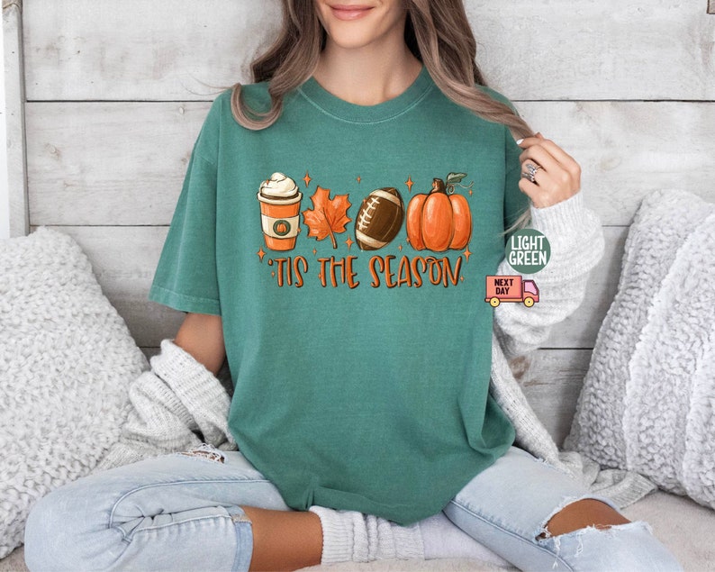 May include: A teal green t-shirt with a graphic of a coffee cup, a football, a pumpkin, and fall leaves. The text "'Tis the season" is printed on the shirt.