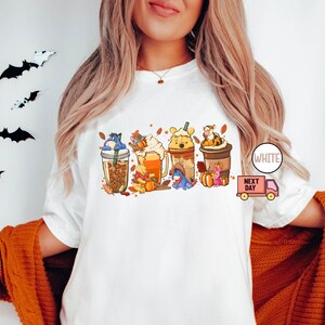 May include: White t-shirt featuring a colorful graphic of fall-themed coffee drinks, each adorned with a different character. The design includes pumpkins, leaves, and the text "WHITE" and "NEXT DAY".