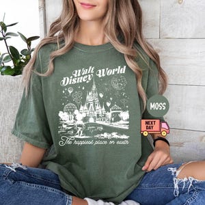 May include: Olive green t-shirt featuring a white graphic of Cinderella's castle, balloons, and fireworks, with the text "Walt Disney World" and "The happiest place on earth". The shirt has short sleeves and a relaxed fit.