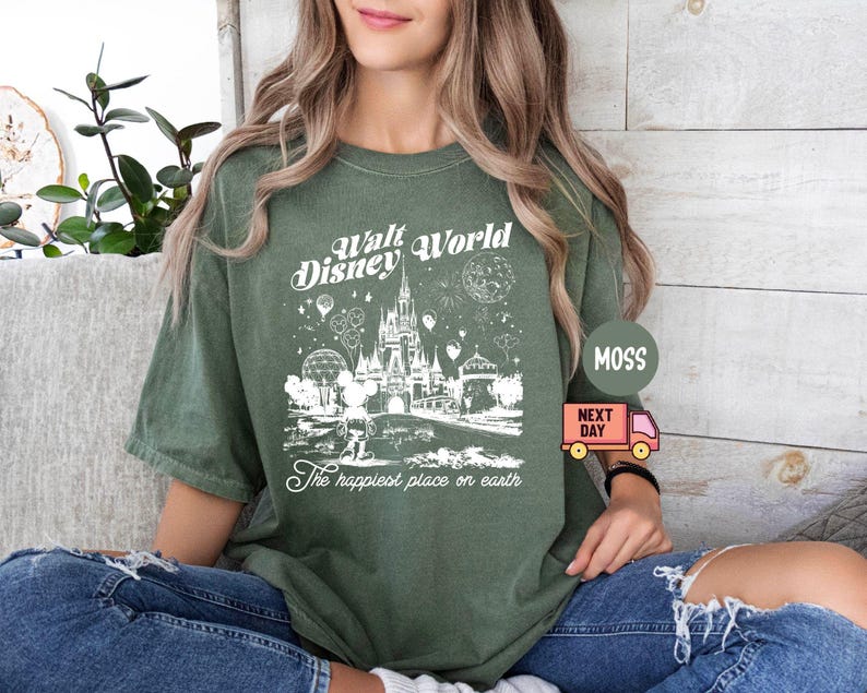 May include: Olive green t-shirt featuring a white graphic of Cinderella's castle, balloons, and fireworks, with the text "Walt Disney World" and "The happiest place on earth". The shirt has short sleeves and a relaxed fit.