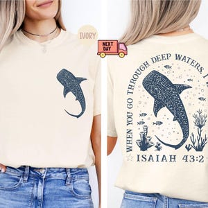 Op de afbeelding: Een cr&egrave;mekleurig T-shirt met een walvishaai-afbeelding en de tekst "When you go through deep waters, I will be with you. Isaiah 43:2" op de achterkant.