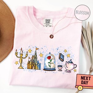 May include: Pink t-shirt with a watercolor design of Beauty and the Beast characters. The design includes a castle, a rose under a glass dome, a teapot, a cup, and a candlestick.