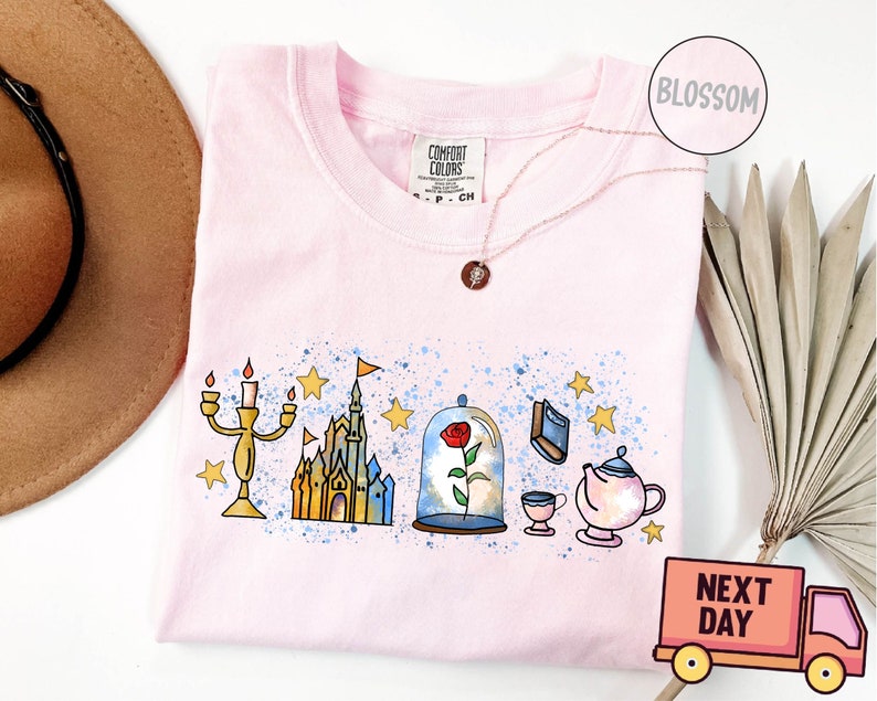 May include: Pink t-shirt with a watercolor design of Beauty and the Beast characters. The design includes a castle, a rose under a glass dome, a teapot, a cup, and a candlestick.