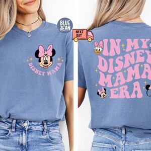 May include: Blue jean t-shirt featuring a Minnie Mouse graphic with a pink bow and the text "Disney Mama" on the front. The back of the shirt has the text "In My Disney Mama Era" in pink, along with additional Disney-themed graphics.