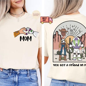 Pu&ograve; includere: Una t-shirt color avorio con una grafica frontale di due mani che formano un cuore sopra la scritta MOM. La grafica sul retro mostra una scena nel deserto con due personaggi dei cartoni animati davanti a un sole, con la scritta YOU GOT A FRIEND IN ME.