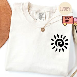 May include: Ivory-colored t-shirt with a black sun graphic. The shirt is a crew neck style and has a small tag on the upper back. A gold necklace with a pendant is draped on the shirt.