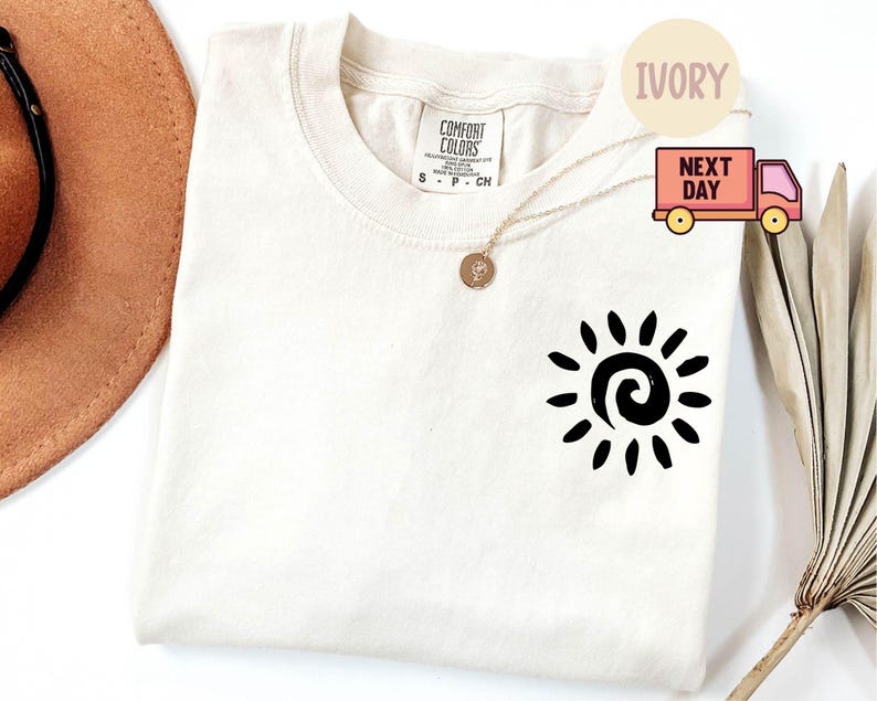 May include: Ivory-colored t-shirt with a black sun graphic. The shirt is a crew neck style and has a small tag on the upper back. A gold necklace with a pendant is draped on the shirt.