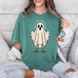 May include: A light green t-shirt featuring a cartoon ghost with the text "Just a Spooky Goose". The ghost has a white body, black eyes, and orange feet. The shirt is a comfortable fit.