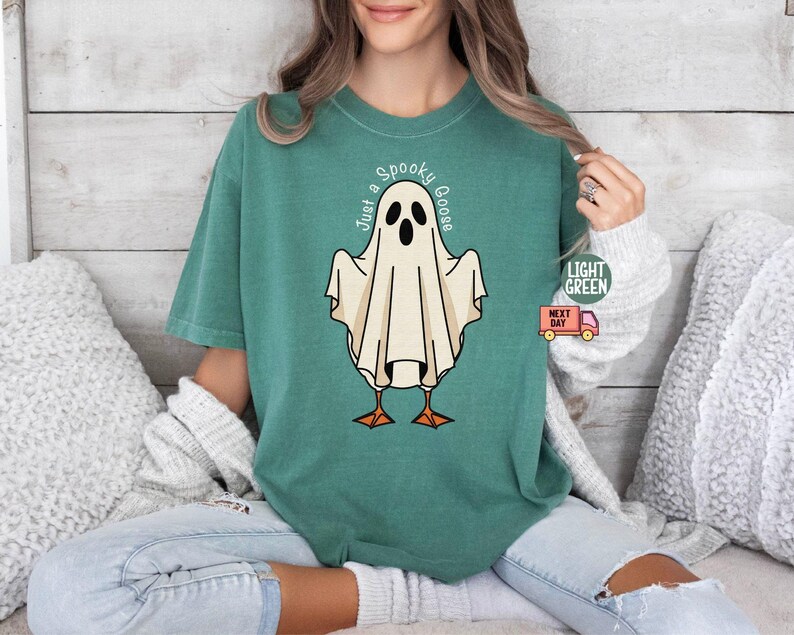 May include: A light green t-shirt featuring a cartoon ghost with the text "Just a Spooky Goose". The ghost has a white body, black eyes, and orange feet. The shirt is a comfortable fit.