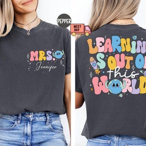 Puede incluir: Dos camisetas grises con un diseño gráfico colorido. La camiseta delantera dice "MRS. Jennifer" con un planeta y un cohete de dibujos animados. La camiseta trasera dice "LEARNING IS OUT OF THIS WORLD" con un planeta y un cohete de dibujos animados.