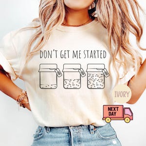 May include: Ivory-colored t-shirt with the text "DON'T GET ME STARTED" above three jars with varying levels of contents. The shirt also features a small pink delivery truck graphic with the words "NEXT DAY".