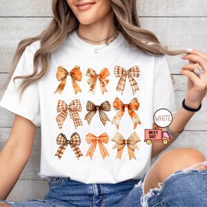 May include: White t-shirt with a design of nine brown and orange bows with plaid patterns. The shirt is on a model wearing blue jeans.