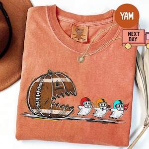 Puede incluir: Camiseta naranja Comfort Colors con una calabaza con forma de balón de fútbol y boca sonriente. Tres figuras de fantasmas con cascos corren hacia la derecha. Se ve un collar de oro.