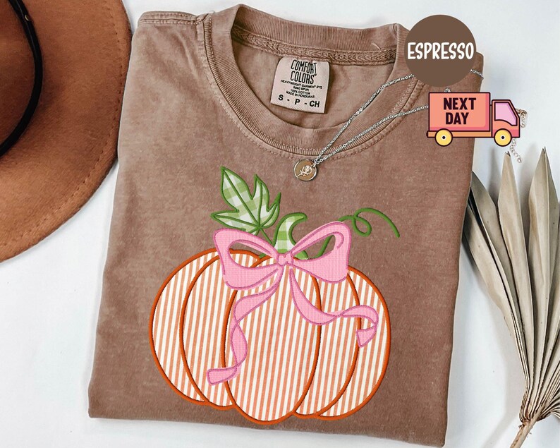 May include: Espresso-coloured Comfort Colors t-shirt featuring a pumpkin design with a pink bow and green leaves. The pumpkin has white and orange stripes. A silver necklace is draped on the shirt.