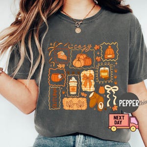 May include: Dark grey t-shirt featuring a collage of autumn-themed illustrations. The design includes pumpkins, a slice of pie, a candle, and the word "cozy". The shirt has a relaxed fit and short sleeves, perfect for fall.