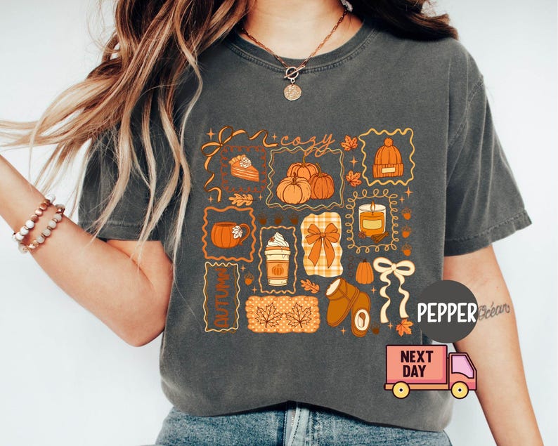 May include: Dark grey t-shirt featuring a collage of autumn-themed illustrations. The design includes pumpkins, a slice of pie, a candle, and the word "cozy". The shirt has a relaxed fit and short sleeves, perfect for fall.