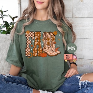 May include: A moss green short-sleeve t-shirt featuring a graphic print of the word FALL in large block letters. The letters are decorated with a brown checkered pattern, cactus silhouettes, and a pair of western boots with floral accents.