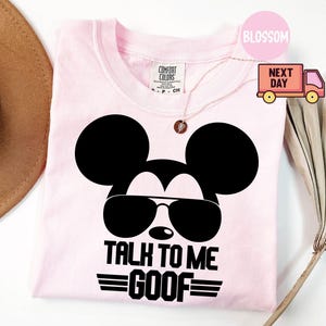 May include: A light pink t-shirt featuring a black graphic of a mouse head wearing sunglasses. Below the graphic, the text reads "TALK TO ME GOOF" in bold black capital letters. The shirt has a Comfort Colors brand label at the inner neckline.