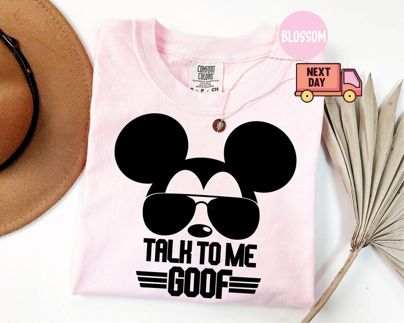 May include: A light pink t-shirt featuring a black graphic of a mouse head wearing sunglasses. Below the graphic, the text reads "TALK TO ME GOOF" in bold black capital letters. The shirt has a Comfort Colors brand label at the inner neckline.