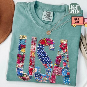 May include: Light green t-shirt with the word "USA" appliqued on the front. The letters are made of colorful floral and star-spangled fabric patches. The shirt has a crew neck and short sleeves.