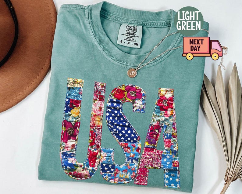 May include: Light green t-shirt with the word "USA" appliqued on the front. The letters are made of colorful floral and star-spangled fabric patches. The shirt has a crew neck and short sleeves.