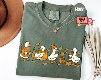 Puede incluir: Camiseta verde musgo Comfort Colors con una ilustración caprichosa de patos con atuendos otoñales. Los patos se representan con calabazas, tazas de café y hojas de otoño. La camiseta tiene cuello redondo y mangas cortas.