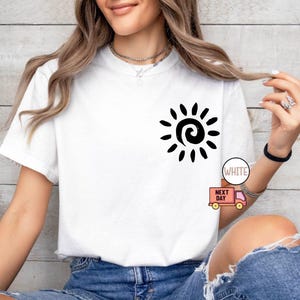 May include: White t-shirt with a black sun graphic on the front. The sun design features a spiral center and radiating lines. The shirt is paired with blue denim shorts. The text "WHITE" is visible on the image.