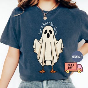 May include: Navy blue t-shirt featuring a cartoon ghost with goose feet and the text "Just a Spooky Goose" in a circular design. The ghost is white with black eyes and mouth. The shirt is a comfortable, casual style.