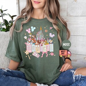 May include: Green t-shirt with a colorful graphic featuring cartoon characters, a castle, and the word "magical." The design includes pastel-colored elements like balloons, bows, and a magic wand. The shirt is a relaxed fit.