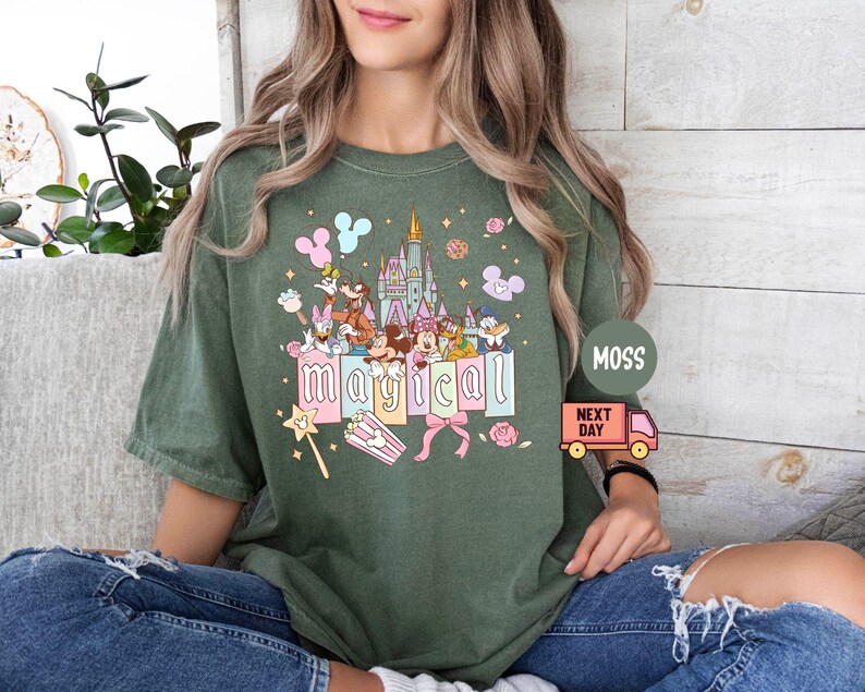 May include: Green t-shirt with a colorful graphic featuring cartoon characters, a castle, and the word "magical." The design includes pastel-colored elements like balloons, bows, and a magic wand. The shirt is a relaxed fit.