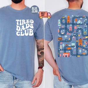 Könnte beinhalten: Blaues Kurzarm-T-Shirt mit dem weißen Schriftzug "Tired Dads Club" auf der Vorderseite. Die Rückseite des Shirts zeigt ein farbenfrohes Straßenkarten-Design mit Autos, Gebäuden und Bäumen. Das Shirt hat eine blaue Grundfarbe.
