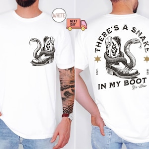 Puede incluir: Camiseta blanca con un diseño en blanco y negro de una bota de vaquero y una serpiente. La parte trasera de la camiseta muestra el texto "THERE'S A SNAKE IN MY BOOT" con "Yee-Haw" debajo. Cuello redondo y mangas cortas.