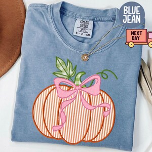 May include: Blue jean coloured t-shirt featuring a pumpkin design. The pumpkin has orange and white stripes, a pink bow, and green leaves. The shirt has a Comfort Colors tag.