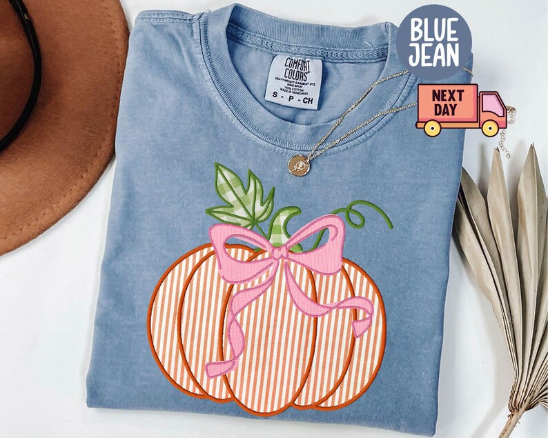 May include: Blue jean coloured t-shirt featuring a pumpkin design. The pumpkin has orange and white stripes, a pink bow, and green leaves. The shirt has a Comfort Colors tag.