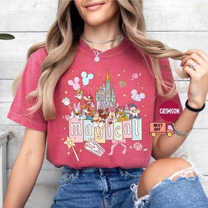 May include: A red t-shirt featuring a colorful graphic of Disney characters, including Mickey and Minnie Mouse, in front of a castle. The word "magical" is spelled out in pastel letters. The shirt has short sleeves.