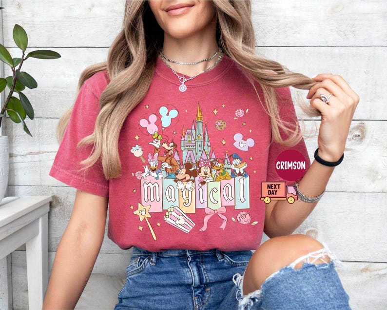 May include: A red t-shirt featuring a colorful graphic of Disney characters, including Mickey and Minnie Mouse, in front of a castle. The word "magical" is spelled out in pastel letters. The shirt has short sleeves.