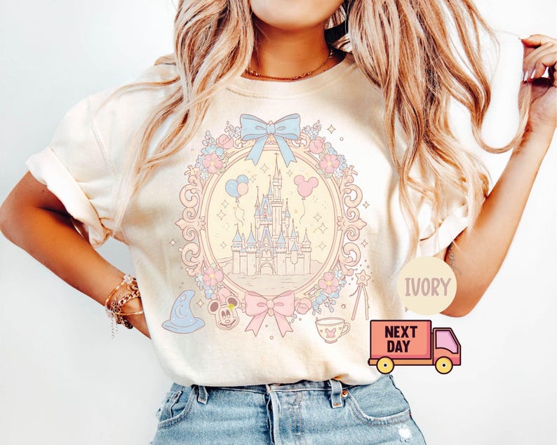 K&ouml;nnte beinhalten: Ein wei&szlig;es T-Shirt mit einer pastellrosa, blauen und gelben Illustration von Schloss Aschenputtel, umgeben von Blumen und einer Schleife. Der Text "IVORY" und "NEXT DAY" ist im Bild sichtbar.
