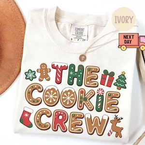 以下が含まれることがあります： アイボリー色のTシャツ。ジンジャーブレッドクッキーのデザインで「THE COOKIE CREW」の文字が描かれています。ジンジャーブレッドマン、雪の結晶、クリスマスツリーのグラフィックが含まれています。金のネックレスが見えます。