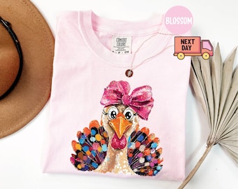 Könnte beinhalten: Hellrosa Comfort Colors T-Shirt mit einem Cartoon-Truthahn-Design. Der Truthahn hat eine rosa Schleife, bunte Federn und große Augen. Eine goldene Halskette ist über dem T-Shirt drapiert.