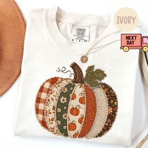 Könnte beinhalten: Ein elfenbeinfarbenes T-Shirt mit Rundhalsausschnitt und einem großen, zentralen Kürbis-Motiv, das aus verschiedenen stoffartigen Flicken in Orange-, Grün-, Braun- und Cremetönen besteht. Das Design zeigt Karos, Blumenmuster und Fischgrät mit einem dunklen Stiel.