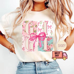 Puede incluir: Una camiseta blanca con un diseño floral rosa y blanco y las letras "Gigi" en una fuente rosa y blanca. Las letras están rodeadas de lazos rosas y diseños florales. La camiseta está disponible en marfil.