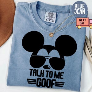 May include: A blue jean colored t-shirt featuring a black graphic of a mouse silhouette wearing sunglasses. The text below the graphic reads TALK TO ME GOOF with horizontal lines. The shirt has a Comfort Colors brand label at the inner neckline.