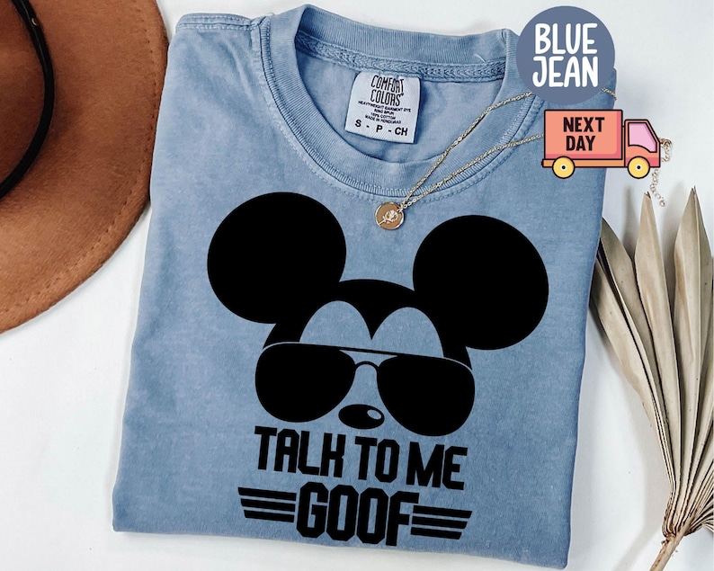 May include: A blue jean colored t-shirt featuring a black graphic of a mouse silhouette wearing sunglasses. The text below the graphic reads TALK TO ME GOOF with horizontal lines. The shirt has a Comfort Colors brand label at the inner neckline.