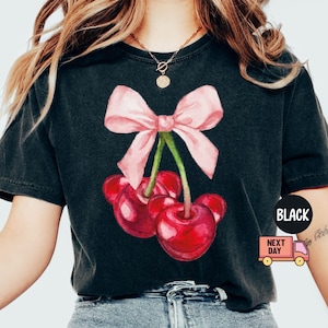 Puede incluir: Camiseta negra con una ilustraci&oacute;n de acuarela de cerezas y un lazo rosa. Las cerezas son de un rojo vibrante y el lazo es rosa claro. La camiseta tiene cuello redondo y mangas cortas.