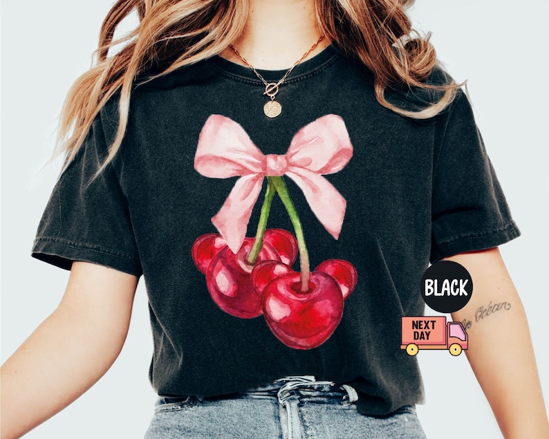 Puede incluir: Camiseta negra con una ilustraci&oacute;n de acuarela de cerezas y un lazo rosa. Las cerezas son de un rojo vibrante y el lazo es rosa claro. La camiseta tiene cuello redondo y mangas cortas.