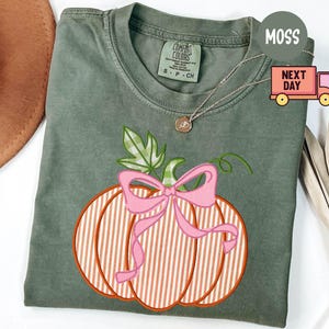 May include: A moss green t-shirt with an embroidered pumpkin design. The pumpkin has orange and white stripes, a pink bow, and green leaves. A gold necklace is visible. The shirt is a Comfort Colors brand.