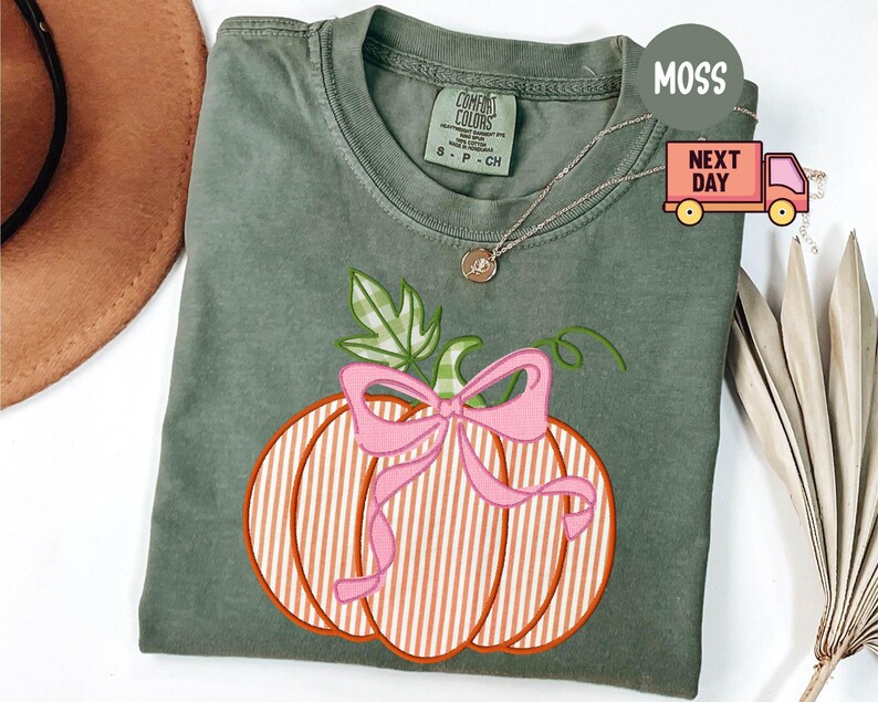 May include: A moss green t-shirt with an embroidered pumpkin design. The pumpkin has orange and white stripes, a pink bow, and green leaves. A gold necklace is visible. The shirt is a Comfort Colors brand.