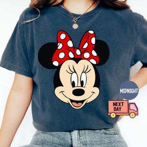 Może przedstawiać: Koszulka w ciemnoniebieskim kolorze z nadrukiem kresk&oacute;wkowej postaci Minnie Mouse. Minnie Mouse ma na sobie czerwoną kokardkę w białe kropki i szeroki uśmiech.